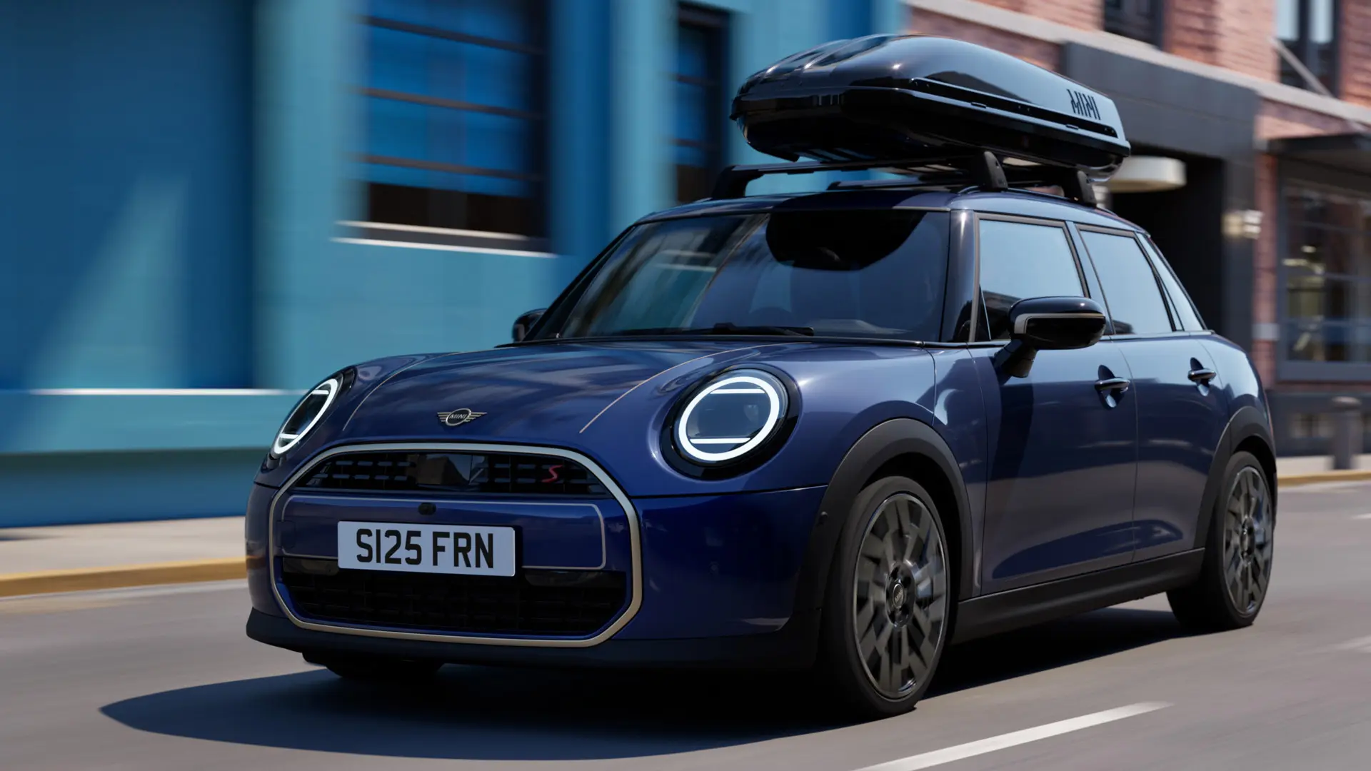 Three quarter front view of MINI Aceman in Indigo Sunset Blue equipped with MINI Roof Rack, carrying a MINI Roof Box 320. 