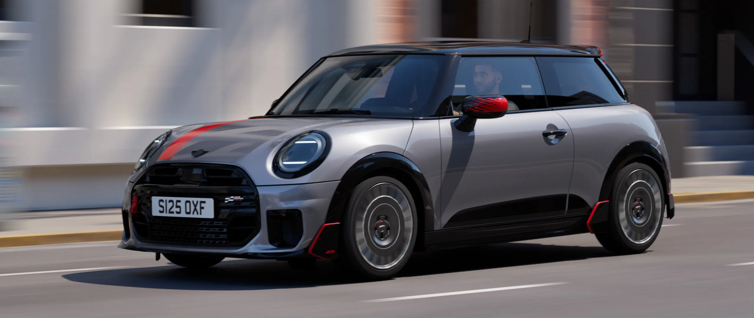 MINI Cooper 3-door in Melting Silver with John Cooper Works mirror cap and bonnet decal in Chili Red, roof spoiler and winglet in Black, driving along city street – front-side view.