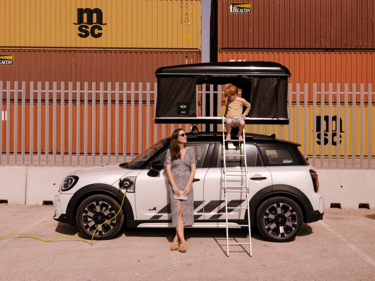 The MINI Countryman is parked in front of shipping containers and is charged at a charging station. Pepe is sitting in the roof tent, his mother is standing in front of the car.